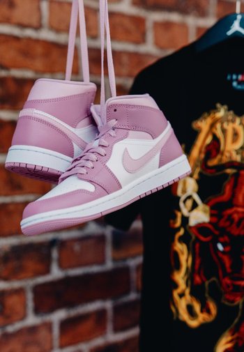 Baskets Nike Air Jordan roses et blanches suspendues par leurs lacets devant un mur de briques et un t-shirt graphique noir.