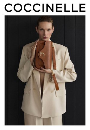 Bolso de cuero marrón con acabado texturizado, herrajes de color dorado y correa ajustable, sostenido por una persona con una blazer beige contra un fondo oscuro.