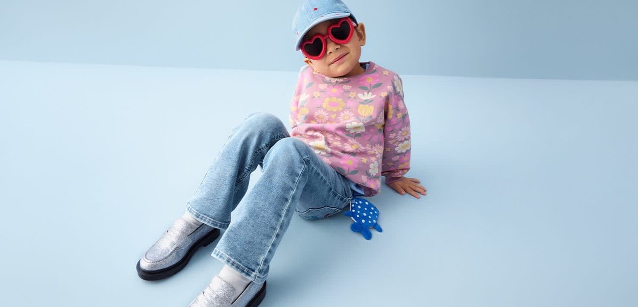 Barn iført røde hjerteformede solbriller, blomsterstriktrøje, blå jeans og kasket sidder på lyseblåt gulv med blå prikker legetøj i nærheden.