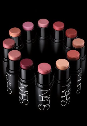 Twelve NARS stick blushes in various colors, including pink, peach, and red, in black cylindrical containers with a glossy finish.
