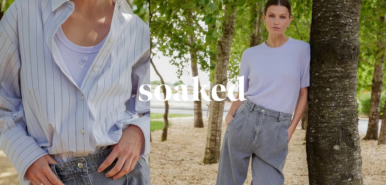 Donna con maglietta azzurra casual e jeans a vita alta, appoggiata a un albero in un ambiente boschivo all'aperto, con le mani nelle tasche.