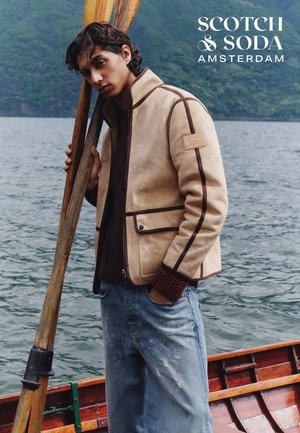 Beige Wildlederjacke mit braunen Akzenten und einem Logo-Patch. Getragen über einem dunklen Pullover und kombiniert mit ausgewaschenen blauen Jeans. Holzhaken in der Hand.