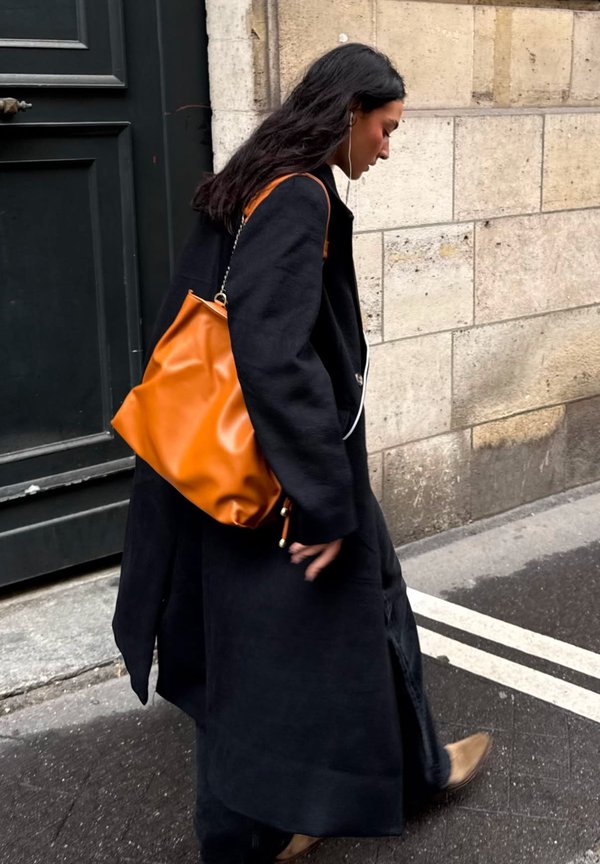 Abrigo negro de mangas anchas y corte largo; lleva un bolso de cuero naranja de forma blanda; caminando por una calle de la ciudad.