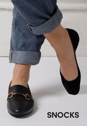 En fod iført en sort loafer med gulddetalje, den anden iført en sort flad sko, begge kombineret med blå opkørte jeans på en lys baggrund.