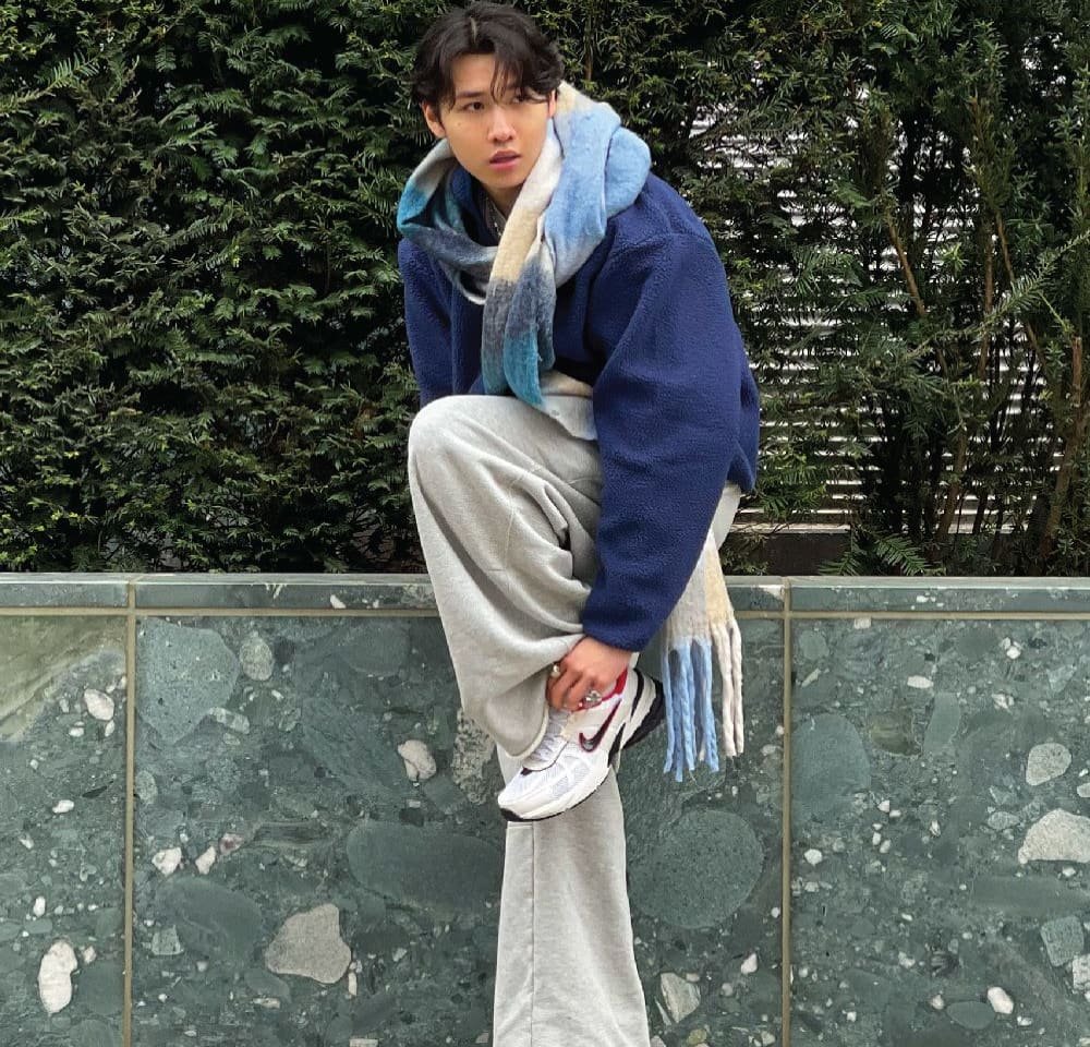 Blue fleece jacket, light gray sweatpants, and white sneakers. Scarf with blue and beige hues. Posed on a stone ledge with greenery in background.