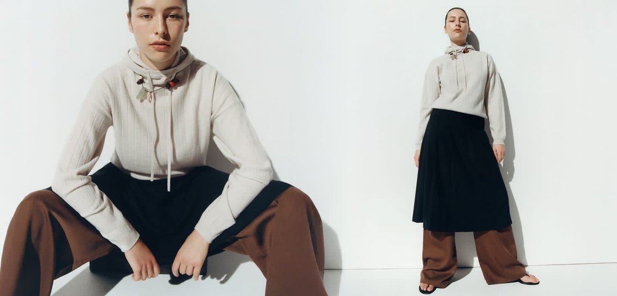 Young woman wearing a cream hoodie, black skirt, and brown wide pants, posing against a white wall in seated and standing positions.