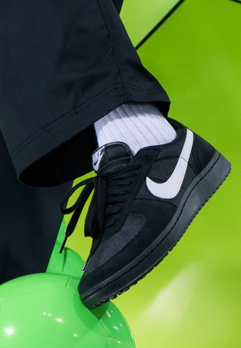 Schwarzer Nike Sneaker mit grauen Akzenten, weißem Swoosh, strukturiertem Stoff und Wildlederelementen, kombiniert mit weißen gestreiften Socken, die auf einem grünen Ball ruhen.