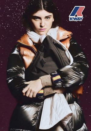 Schwarz-orangefarbene Steppjacke mit glänzender Textur und kontrastierender weißer Innenschicht. Das Model hält eine dunkle Tasche und zeigt mehrfarbige Manschettenakzente.