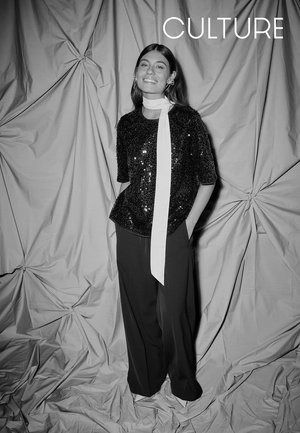 Schwarzes Paillettenoberteil mit kurzen Ärmeln und weißem Schal, kombiniert mit weiten schwarzen Hosen. Der Hintergrund zeigt drapierte graue Stoffe.