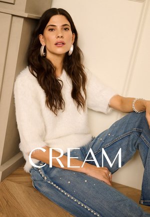 Pull blanc moelleux, jeans en denim bleu avec des accents en perles le long des coutures, modèle assis contre un mur, sol en bois doux.