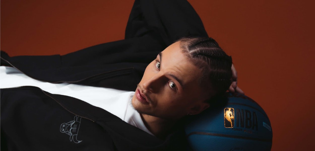 Giovane con capelli intrecciati disteso con la testa appoggiata su un pallone da basket NBA blu su sfondo marrone.