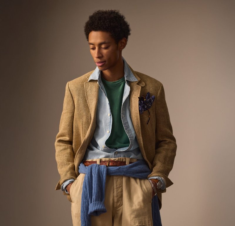 Blazer en chevrons couleur tan, superposé sur une chemise en denim et un t-shirt vert. Pull bleu noué à la taille, complété par un pantalon beige.