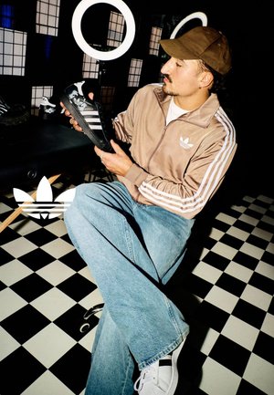 Black Adidas Superstar sneaker with white accents, held by a person in a beige jacket. Black and white checkered floor in the background.