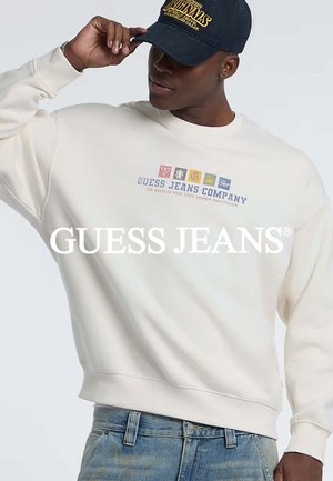 Cremefarbener Sweater mit Rippbündchen, der ein zentrales Logo mit bunten Emblemen und dem Text "GUESS JEANS COMPANY" zeigt, kombiniert mit blauen Jeans.