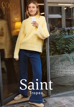 Gelber, strukturierter, übergroßer Pullover mit weiten Ärmeln, kombiniert mit dunkelblauen, gestreiften, weit geschnittenen Hosen und beigen Slippern. Hält eine Kaffeetasse in der Hand.