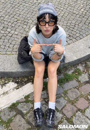 Person med solbriller, en stribet hovedbund, grå sweater og sorte sneakers, siddende på en stenkrans med brostensbelagt jord.