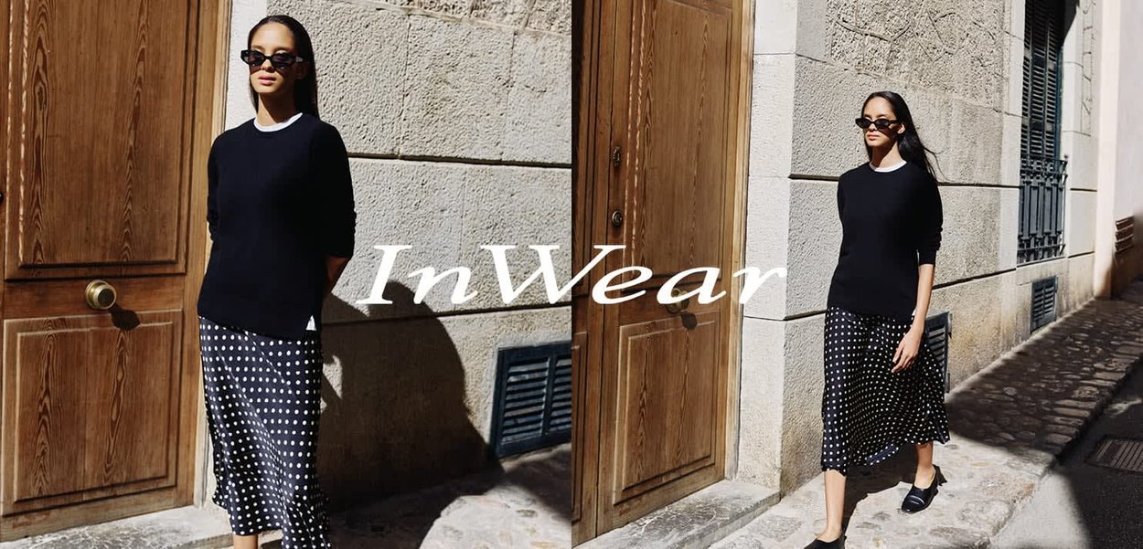 Schwarzer Pullover mit langen Ärmeln, gepunkteter Midirock, dunkle Sonnenbrille und flache Schuhe, stehend vor einer strukturierten Steinmauer.