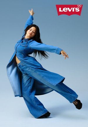 Denimoutfit bestaande uit een lange, open jas met knopen en wijde spijkerbroeken, beide in verschillende tinten blauw, tegen een lichtblauwe achtergrond.