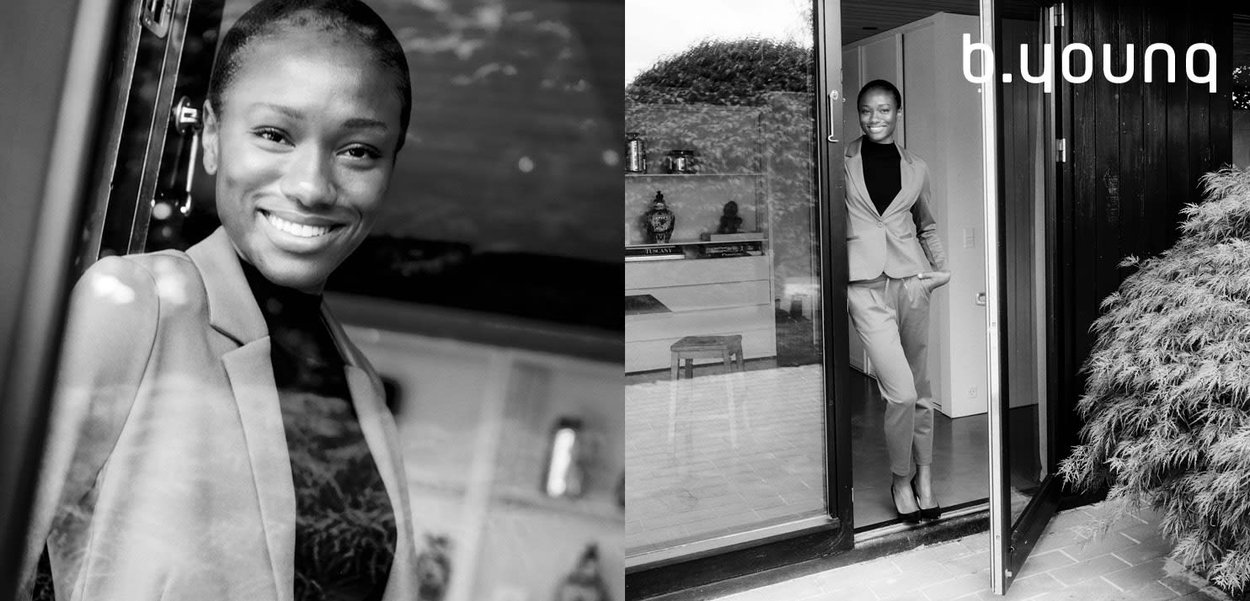 Eine Frau in einem maßgeschneiderten hellgrauen Hosenanzug steht in einer Türöffnung und lächelt. Der Raum ist mit Holzakzenten und Zimmerpflanzen gestaltet.