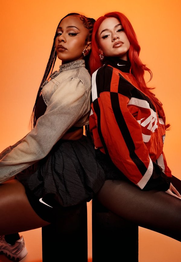 Zwei Models sitzen Rücken an Rücken. Eine trägt eine helle Jeansjacke und einen schwarzen Minirock; die andere trägt eine rot-schwarze Nike-Regenjacke und Leggings.