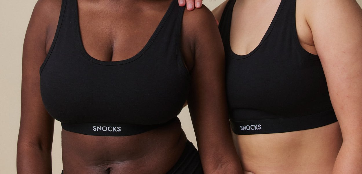 To kvinder iført sorte sports-bh'er med mærket "SNOCKS", en mørk og en lys i huden, stående tæt sammen med en hånd på den andens skulder.