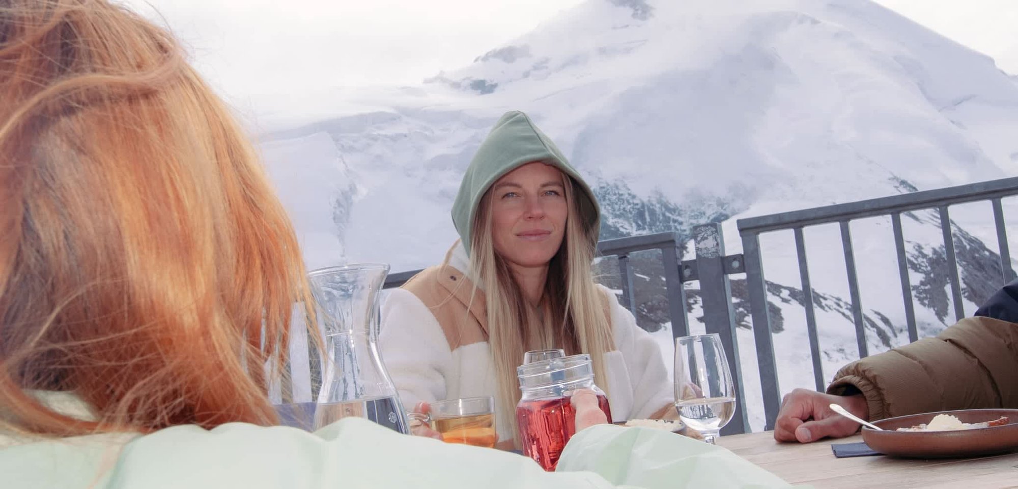 Eine Frau in einem grünen Hoodie und einem cremefarbenen Mantel sitzt an einem Holztisch im Freien. Im Hintergrund sind schneebedeckte Berge und ein bewölkter Himmel zu sehen.