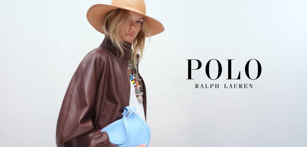 Vrouw in bruin leren jasje en brede hoed die een lichtblauwe handtas vasthoudt, met kleurrijke ketting, tegen een effen lichte achtergrond.