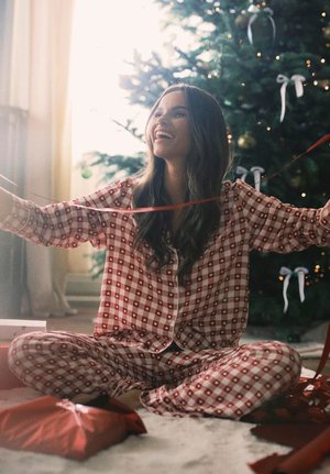 Femme en pyjama à carreaux rouges assise en tailleur près d'un sapin de Noël décoré, tenant un ruban rouge, souriante et heureuse.
