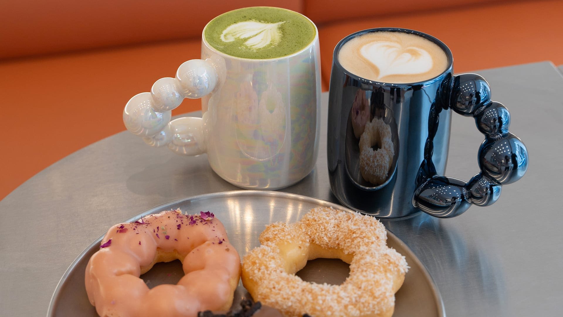 Twee unieke koffiemokken: de ene glanzend wit met een bubbelfvormige handgreep, de andere glanzend zwart. Drie donuts op een metalen bord: roze, kokosnoot en chocolade.