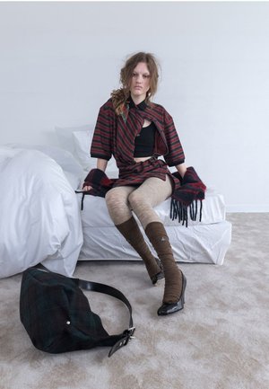 Veste en tartan portée sur un top noir, associée à des chaussettes hautes jusqu'aux genoux. Un sac texturé au premier plan. Un arrière-plan avec une literie et un tapis aux teintes neutres.