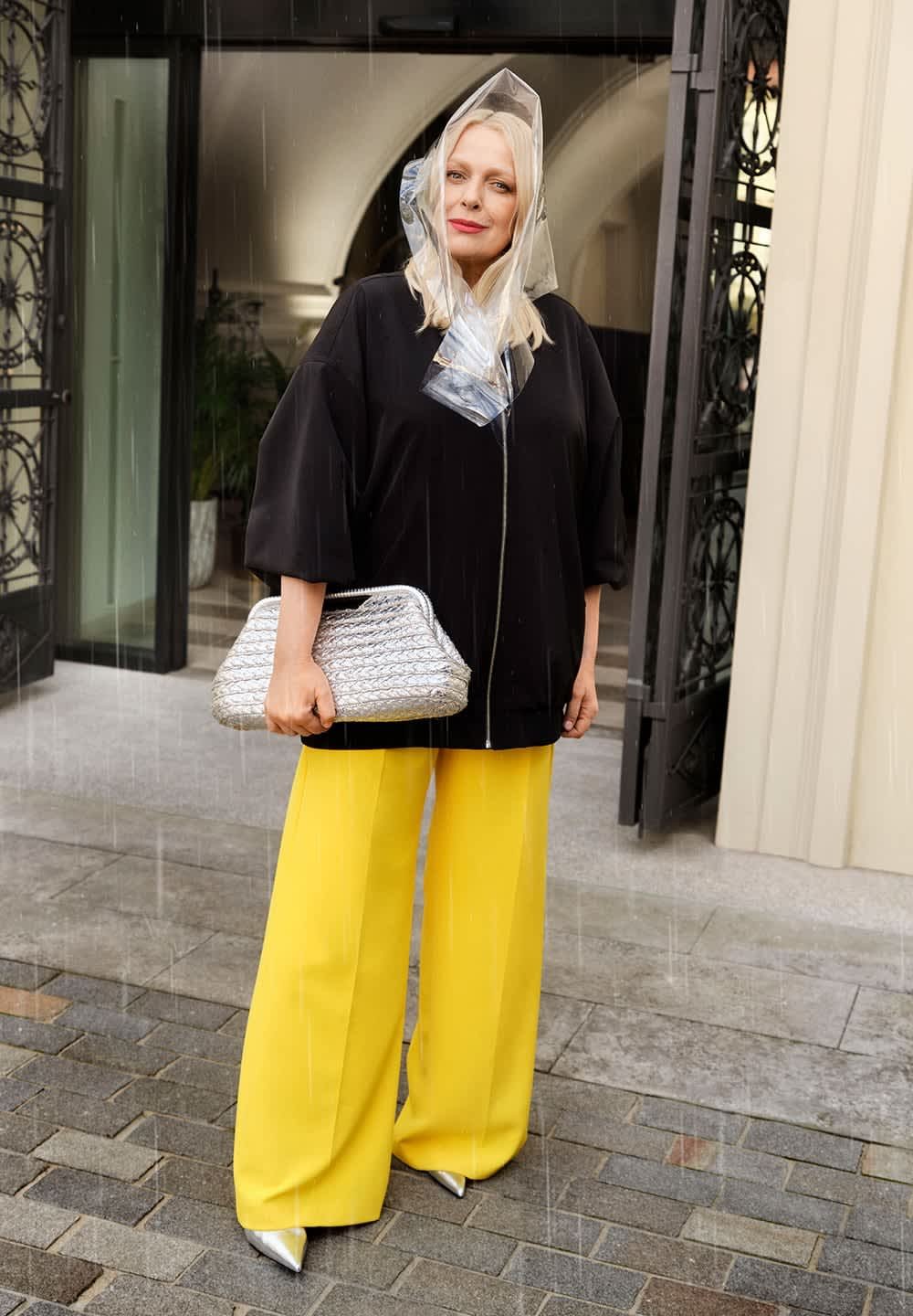 Czarna, oversize kurtka z srebrnym zamkiem, zestawiona z jasnobirkowymi, szerokimi spodniami oraz metaliczno-srebrnymi, szpiczastymi butami. Trzyma teksturowaną srebrną kopertówkę.