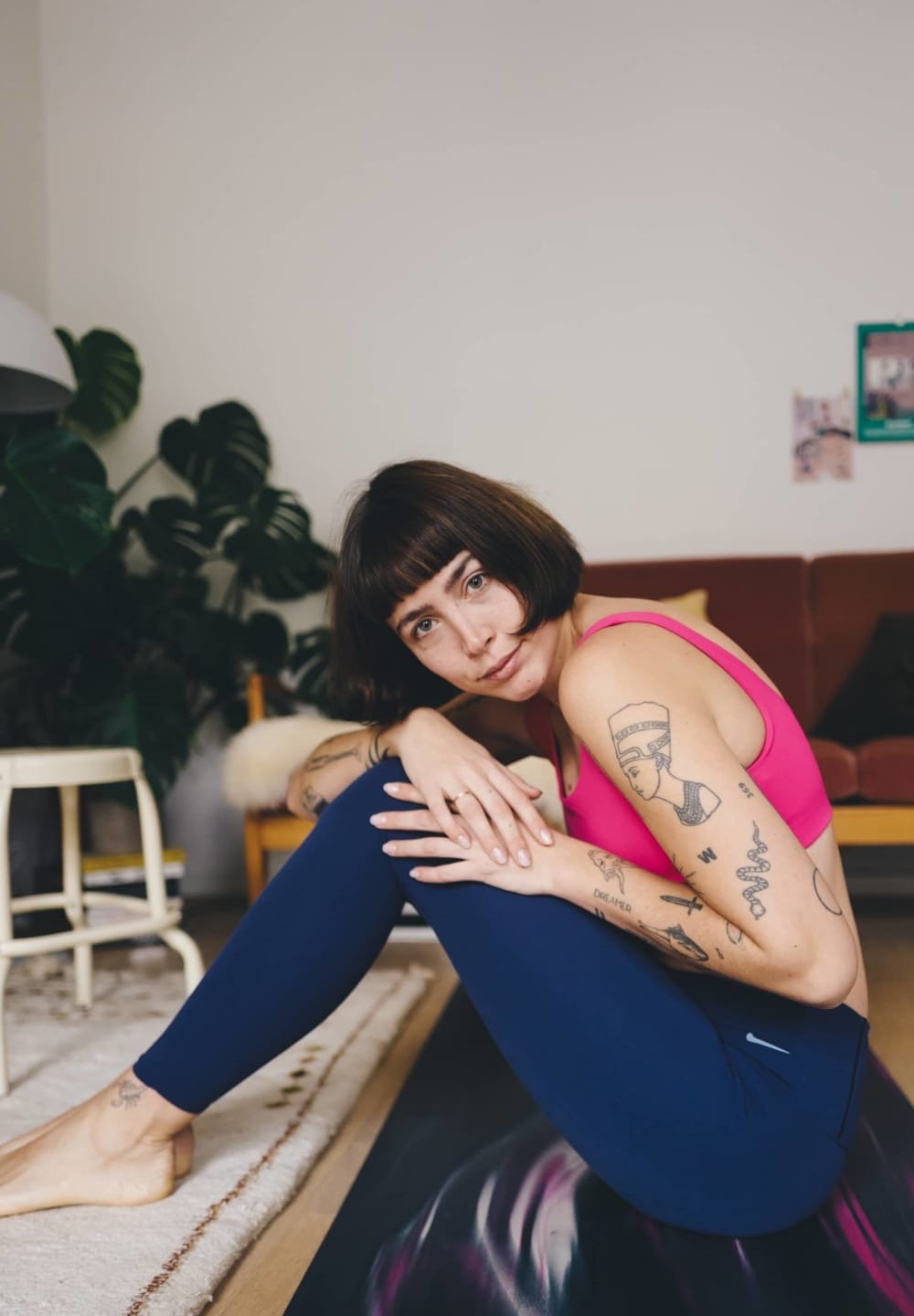 Fitted pink tank top and navy leggings, displaying tattoos. Indoor setting with plants, wooden furniture, and a rug. Casual pose on a mat.