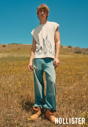 Un jeune homme se tient dans un champ portant un t-shirt blanc sans manches avec un motif de flammes, un jean bleu ample et des bottes de travail beige sous un ciel bleu clair.