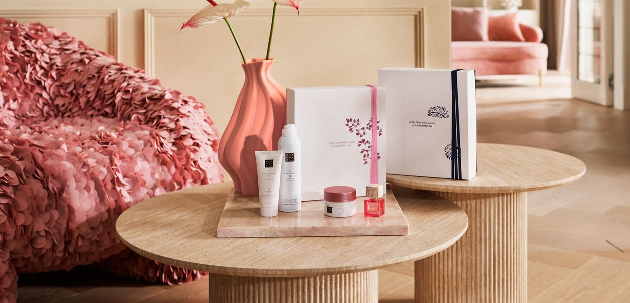 Tables rondes en bois avec des produits de soin de la peau, un vase rose avec des fleurs et un canapé rose texturé dans une pièce confortable et doucement éclairée.