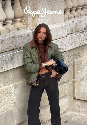 Groene gewatteerde jas met een bruine corduroy kraag, bruin overhemd met knopen, zwarte hoge taille broek en een zwarte handtas tegen een stenen muur.