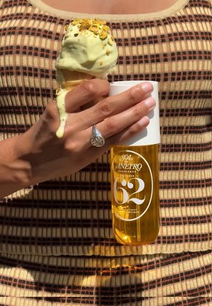 Ręka trzymająca topniejący rożek lodów pistacjowych i bursztynową butelkę mgiełki perfum Sol de Janeiro, ubrana w brązowy, prążkowany top.