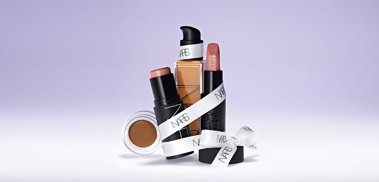 Produits cosmétiques NARS présentés : un flacon de fond de teint transparent, deux rouges à lèvres (rose et nude), un bronzer crème, et un ruban blanc avec le logo.