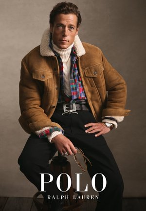 Brun mockaskinnjacka med shearlingkrage, över en rutiga skjorta, vit polotröja och svarta byxor. Har ett bälte med silverspänne och klocka.