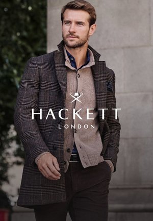 Geruit wollen mantel over een beige gebreide trui, daaronder een marineblauw overhemd, bruine ribfluwelen broek. Voorzien van een pochet en details van een textuurrijk stof.