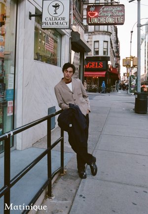 Mann in beigem Blazer, der sich auf einem Geländer vor einer Apotheke auf dem Bürgersteig einer Stadt lehnt, mit Geschäften und Passanten im Hintergrund.