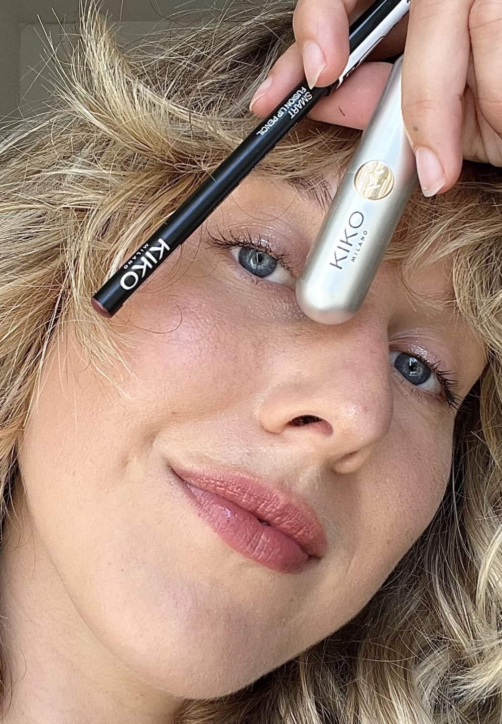 Mascara argent et eyeliner noir de KIKO Milano, tubes lisses, accents de logo, présentés sur un fond clair avec des cheveux ondulés doux.