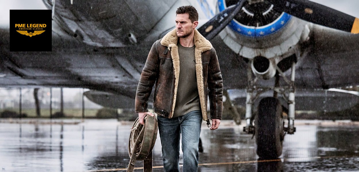 Veste en cuir marron avec doublure en fourrure, pull gris, jeans bleus, bottes noires. Portant un sac à bandoulière, debout sur une piste mouillée près d'un avion.