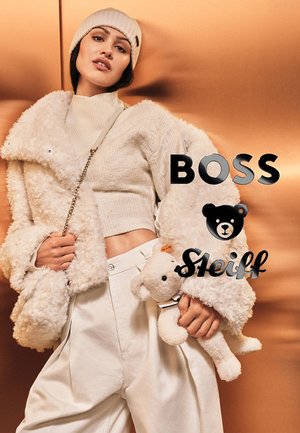 Flauschige weiße Jacke mit plüschiger Textur, kombiniert mit einem leichten Pullover und weit geschnittenen cremefarbenen Hosen. Hält einen kleinen Teddybären.