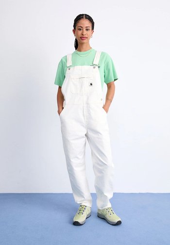 Witte denim schort met verstelbare banden, een voorvak en een relaxte pasvorm, gecombineerd met een lichtgroen T-shirt en grijze sneakers.