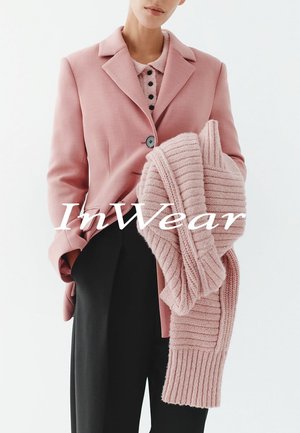 Rosa Blazer mit Reverskragen und vorderer Knopfleiste, kombiniert mit einem hellrosa gestrickten Pullover mit gerippter Struktur. Schwarze Hose.