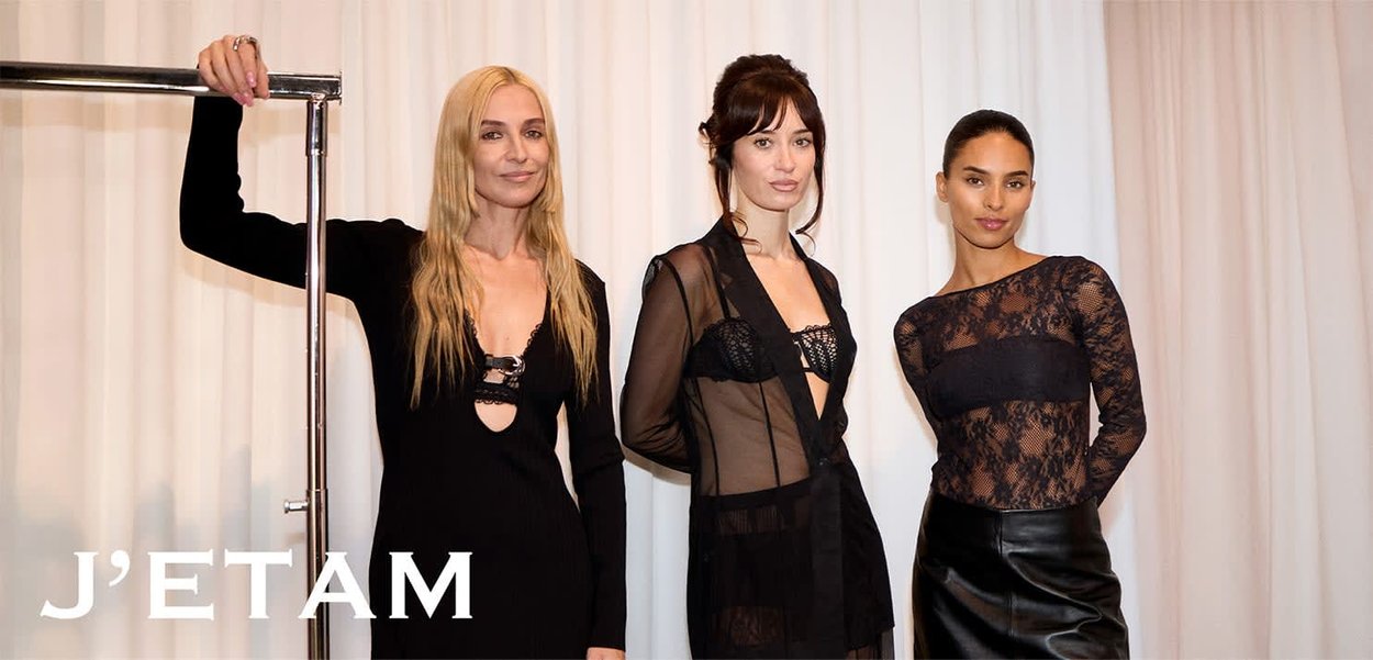 Drei Frauen stehen in schwarzen, transparenten und figurnahen Outfits vor weißen Vorhängen, wobei eine von ihnen auf der linken Seite ein Metallkleidergestell hält.