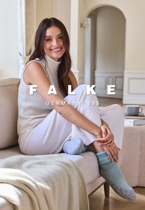 Hellgrauer, gerippter Strickrollkragen und weiße Hosen mit weitem Bein, dazu strukturierte blaue und graue Knöchelsocken, sitzend auf einem beigen Sofa.