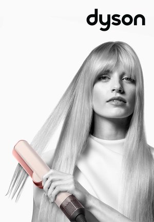 Dyson hair straightener in pink and white, held in a hand, features a sleek design and glossy finish, set against a light background.