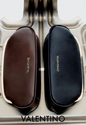 Zwei Valentino-Ledertaschen, eine braun und eine schwarz, nebeneinander auf einer reflektierenden Metalloberfläche mit Valentino-Logo darunter präsentiert.