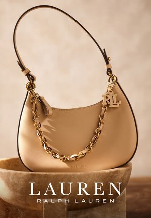 Beige Leder-Schultertasche mit goldener Kettenriemen und RL-Goldinitial-Anhänger, präsentiert auf einem Steinsockel vor neutralem Hintergrund.